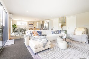 A modern living room with a sofa, ottoman, and coffee table, a dining area with a table and chairs, and a kitchen with a refrigerator and stove.