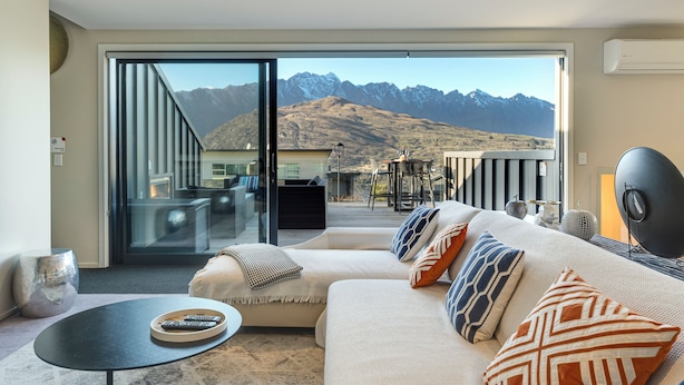 A modern living room with a large sofa, a small round table, and a glass door leading to a balcony with a mountain view.