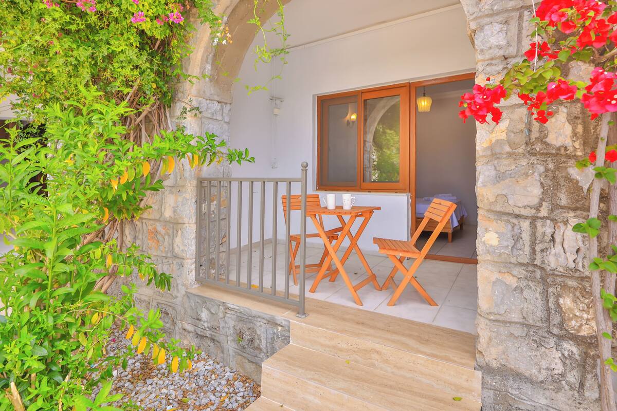 A stone archway leading to a balcony with a table and chairs, surrounded by greenery and flowers.