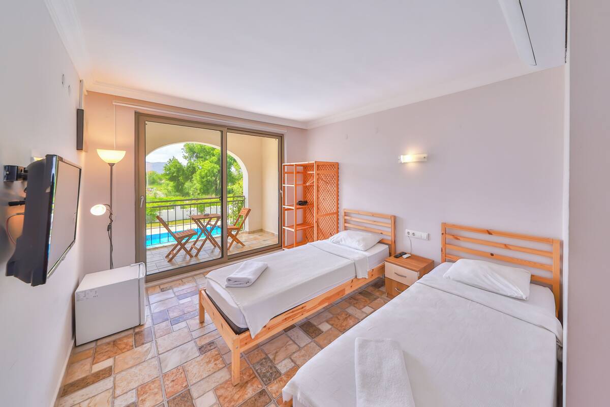 A bedroom with two beds, a TV, a desk, a chair, a lamp, and a sliding glass door leading to a balcony with a pool.