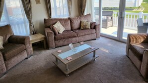 A cozy living room with a brown sofa, two armchairs, a glass coffee table, and a sliding glass door leading to an outdoor patio. - 2br-sleeps6-fireplace-parking-patio-seaside (Clacton-on-Sea)