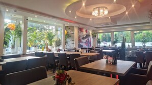 A restaurant with multiple tables and chairs, large windows with plants, and a decorative ceiling light. - SILONCE Hotels & Restaurant (Radevormwald)