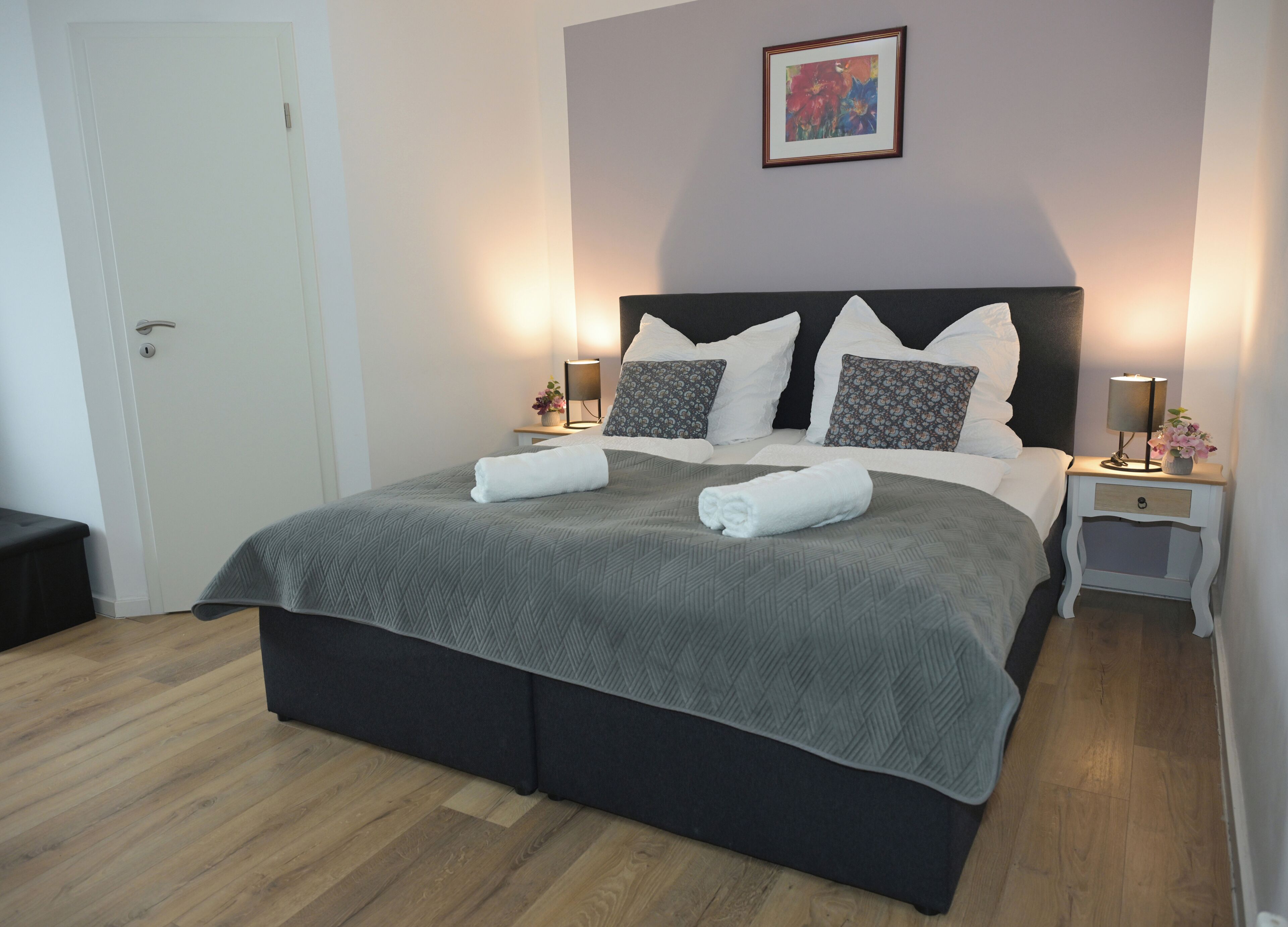 A neatly made bed with a grey bedspread and two white towels, a bedside table with a lamp, a framed picture on the wall, and a door with a handle.