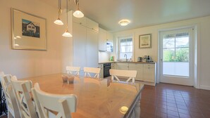 A spacious dining area with a large wooden table and white chairs, a kitchen with white cabinets, and a window with a view of greenery. - House (Wenningstedt)