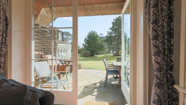 A room with a sliding glass door leading to an outdoor patio area with a stone floor and wooden furniture. - House (Wenningstedt)