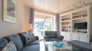 A living room with a sofa, armchair, and glass coffee table. There is a bookshelf with books and a flat-screen TV. - House (Wenningstedt)