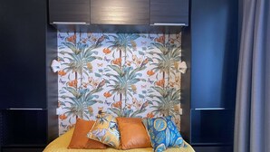 A bed with a textured yellow cover, two pillows with tropical patterns, and a floral wallpaper with birds and leaves.