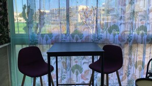 A room with a table and two chairs, a window with a view of buildings, and a curtain with a tropical pattern.