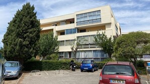 A multi-story building with a flat facade, large windows, and balconies. There are cars parked in front, and a tree is visible to the left.
