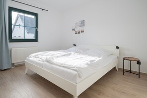 A bedroom with a white bed, a small wooden stool, a window with a view of buildings, and a radiator.
