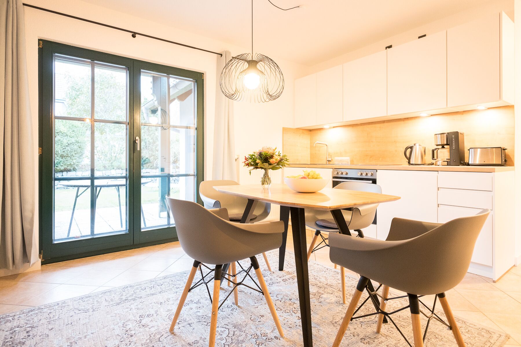 A modern kitchen with white cabinets, a dining table with four chairs, and a large window with a view of the outdoors.