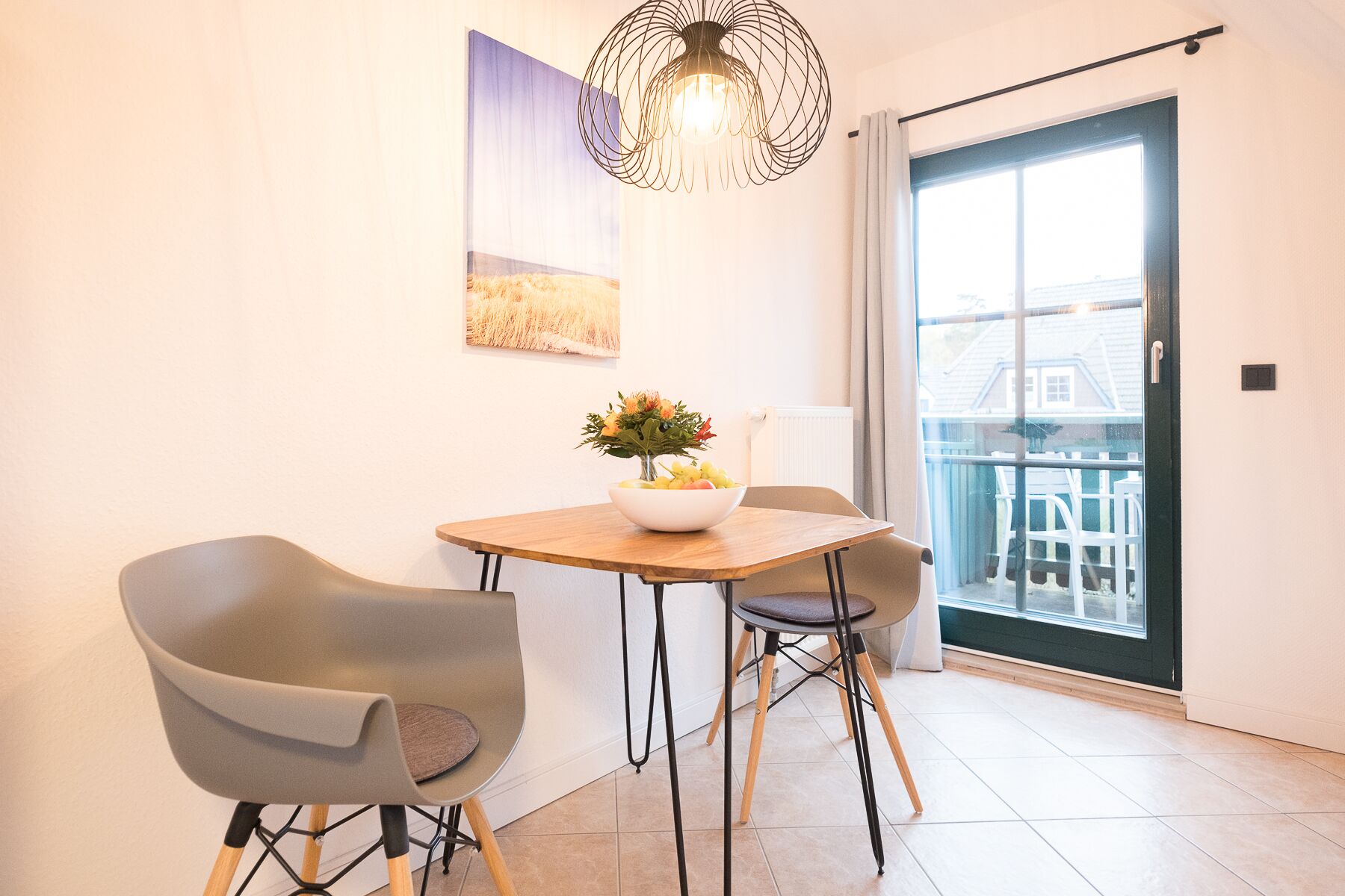 A dining area with a round wooden table, two grey chairs, a vase with flowers, and a hanging light fixture.