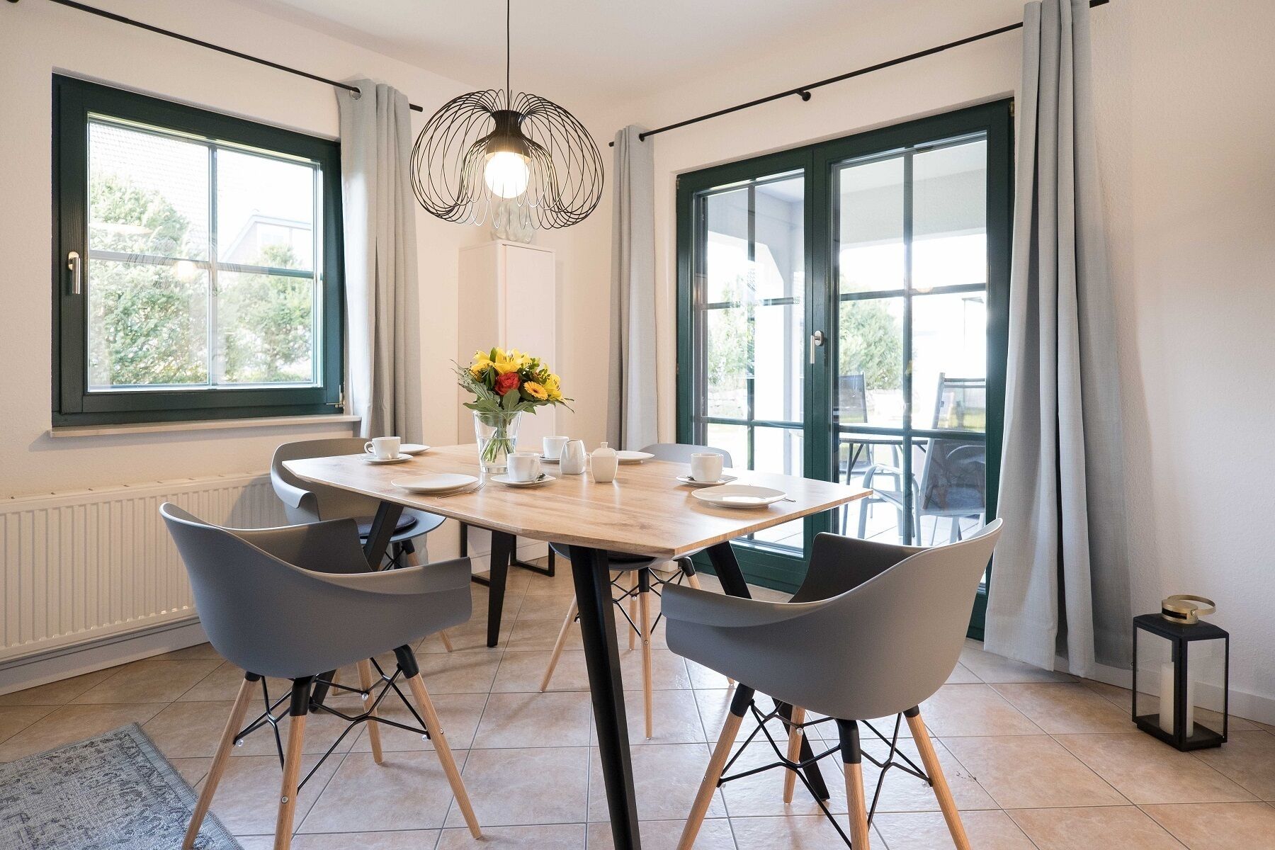 Un coin repas avec une table en bois, quatre chaises grises, un vase avec des fleurs jaunes et un luminaire suspendu.