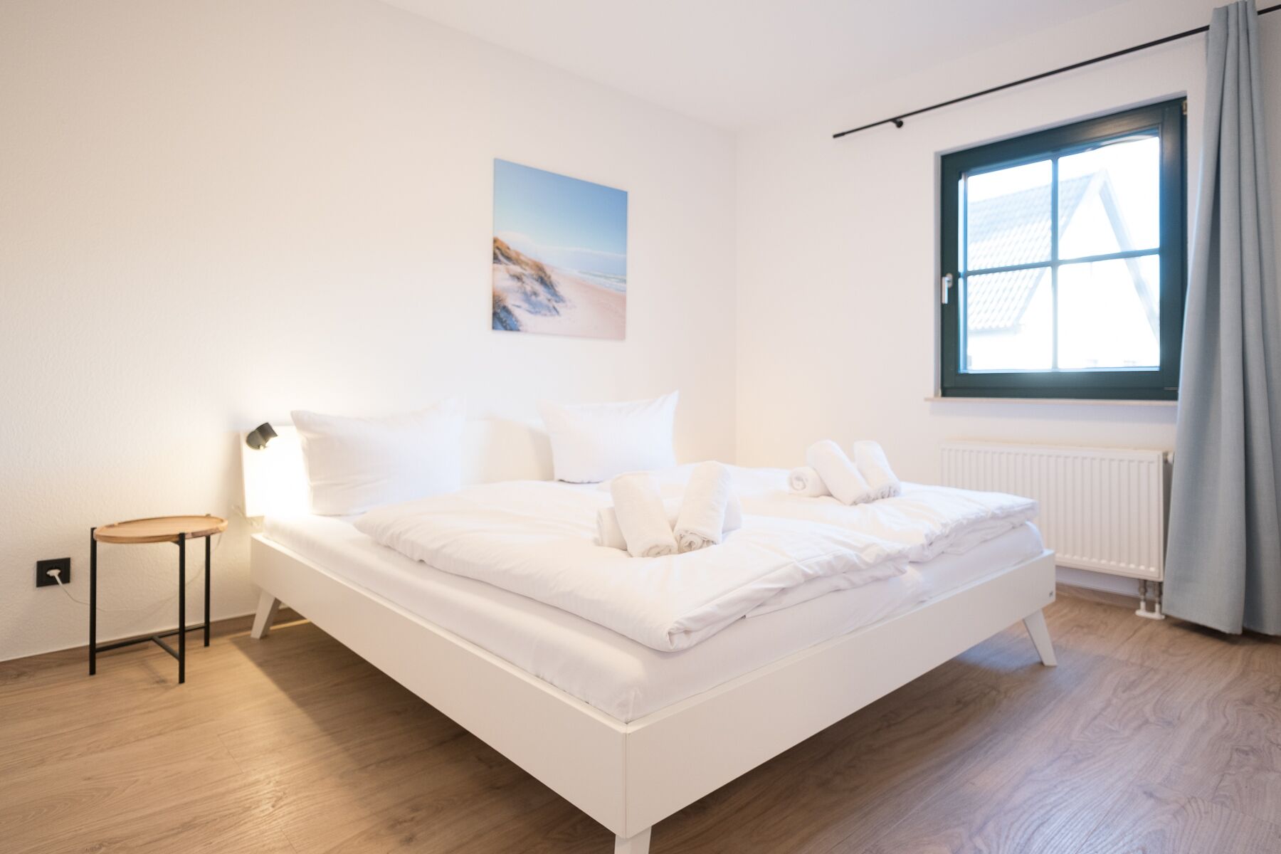 Un lit bien fait, avec du linge de lit blanc, une petite table de chevet, une lampe fixée au mur et une fenêtre avec des rideaux.