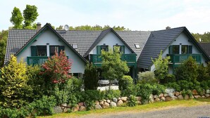 Una hilera de casas blancas con contraventanas verdes y techos grises, rodeadas de vegetación y un muro de piedra.