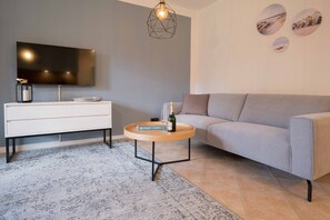 A modern living room with a grey sofa, a round wooden coffee table, a white dresser, and a flat-screen TV mounted on the wall.