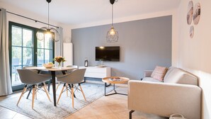 Una sala de estar moderna con un sofá gris, un comedor con mesa y sillas, un televisor montado en la pared y un ventanal con cortinas.