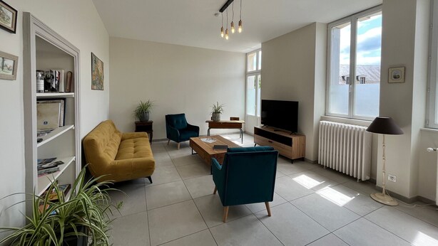 Ein modernes Wohnzimmer mit einer Couch, zwei Sesseln, einem Couchtisch und einem Fernseher. Ücht Blumentöpfen und einem Bücherregal.