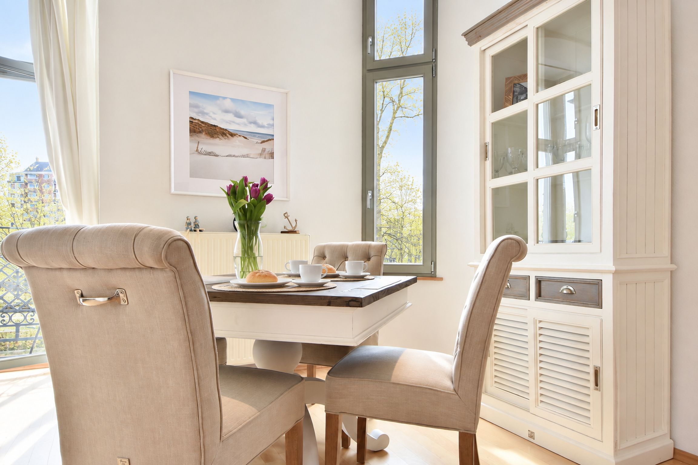 A dining area with a table set for two, chairs, a vase of flowers, and a window with a view of the outside.