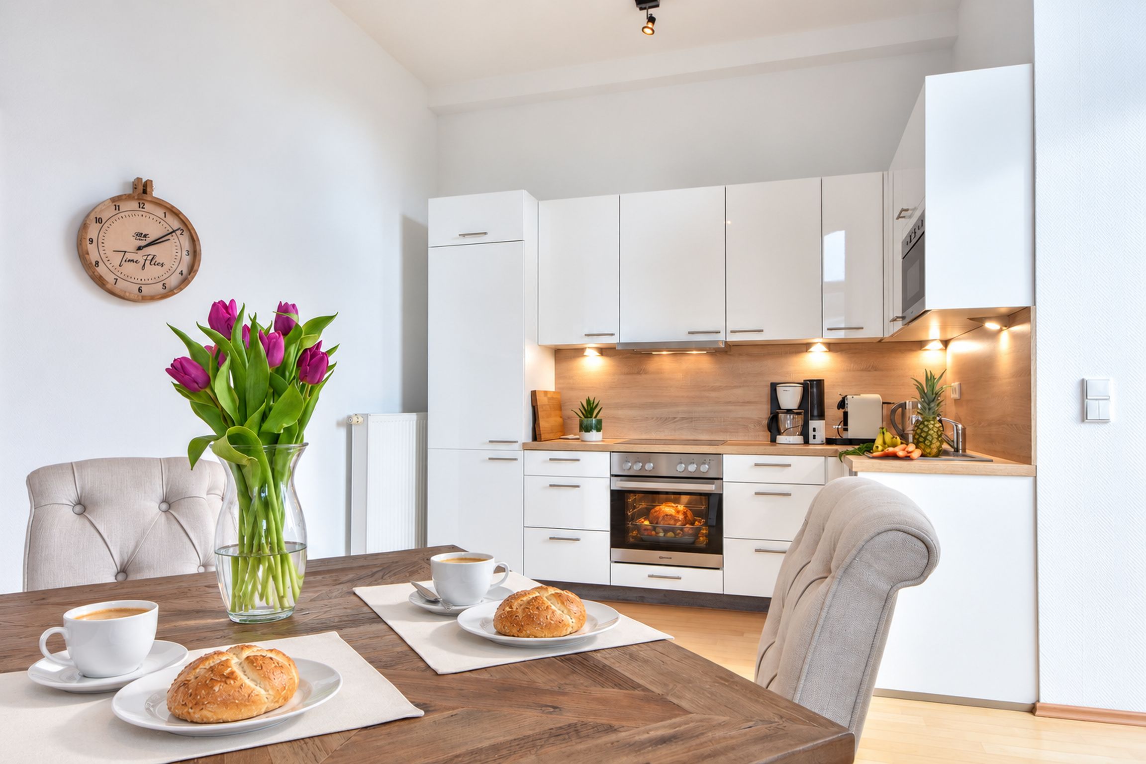 A modern kitchen with white cabinets, a built-in oven, and a dining area with a wooden table set for two.