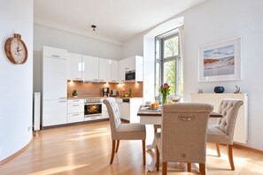 Une cuisine moderne avec des meubles blancs, un coin repas avec deux chaises, une table où sont disposés des aliments pour le petit-déjeuner et une grande fenêtre offrant une belle vue.