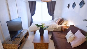 A room with a TV on a glass table, a bed with white bedding, a brown sofa, and a small table with a vase of flowers. - Mishuku Onna102 (Onna)