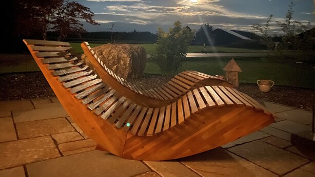 A wooden bench with a curved backrest, set on a paved patio with a view of a landscape and a large rock in the background. - Cozy holiday chalet with top facilities - Hausen on the ground floor - near Chiemsee (Pittenhart)