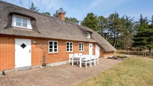 Una casa de ladrillo con techo de paja, una puerta blanca y un pequeño jardín con mesa y sillas.
