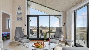 Una sala de estar moderna con ventanas amplias, dos sillones grises, una mesa de centro redonda con aperitivos y vistas a un paisaje lejano.