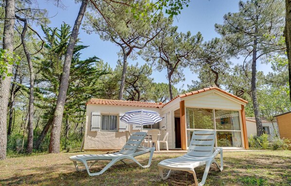 A small, single-story building with a tiled roof, surrounded by tall pine trees. There are two lounge chairs and a striped umbrella in the foreground.
