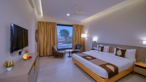 Habitación de hotel con cama, escritorio con televisor, una silla, una mesita con fruta y ventana con cortinas.