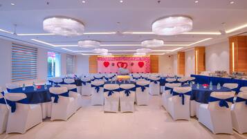 A banquet hall with round tables, white chairs with blue sashes, chandeliers, and a stage with a red backdrop.