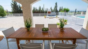 Un espace extérieur abrité, aménagé pour les repas, avec une table et des chaises en bois, entouré de plantes en pot et offrant une vue sur une piscine et un espace verdoyant.