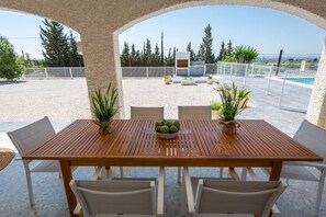 Uma área de refeições coberta ao ar livre, com mesa e cadeiras de madeira, cercada por plantas em vasos e com vista para uma piscina e vegetação.