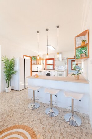 Une cuisine moderne avec des armoires blanches, un comptoir en bois et trois tabourets de bar. On y trouve des luminaires suspendus, une plante en pot et un tableau encadré au mur.