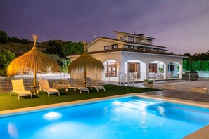 Piscina com espreguiçadeiras e guarda-sóis, uma casa com varanda e céu estrelado.