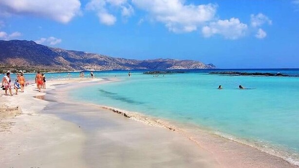 Een strand met helder turquoise water, mensen die zwemmen en over het strand lopen, en bergen op de achtergrond.