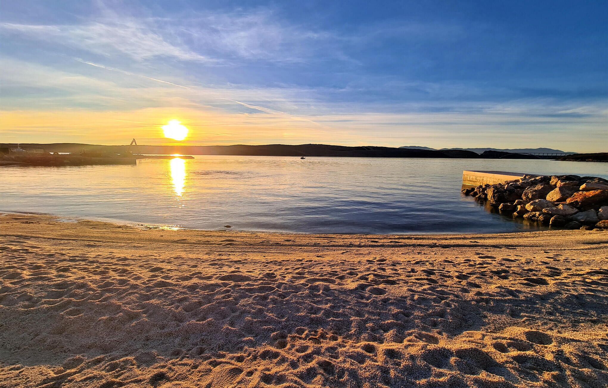A beach with a calm sea, a rocky breakwater, and a sunset in the background.