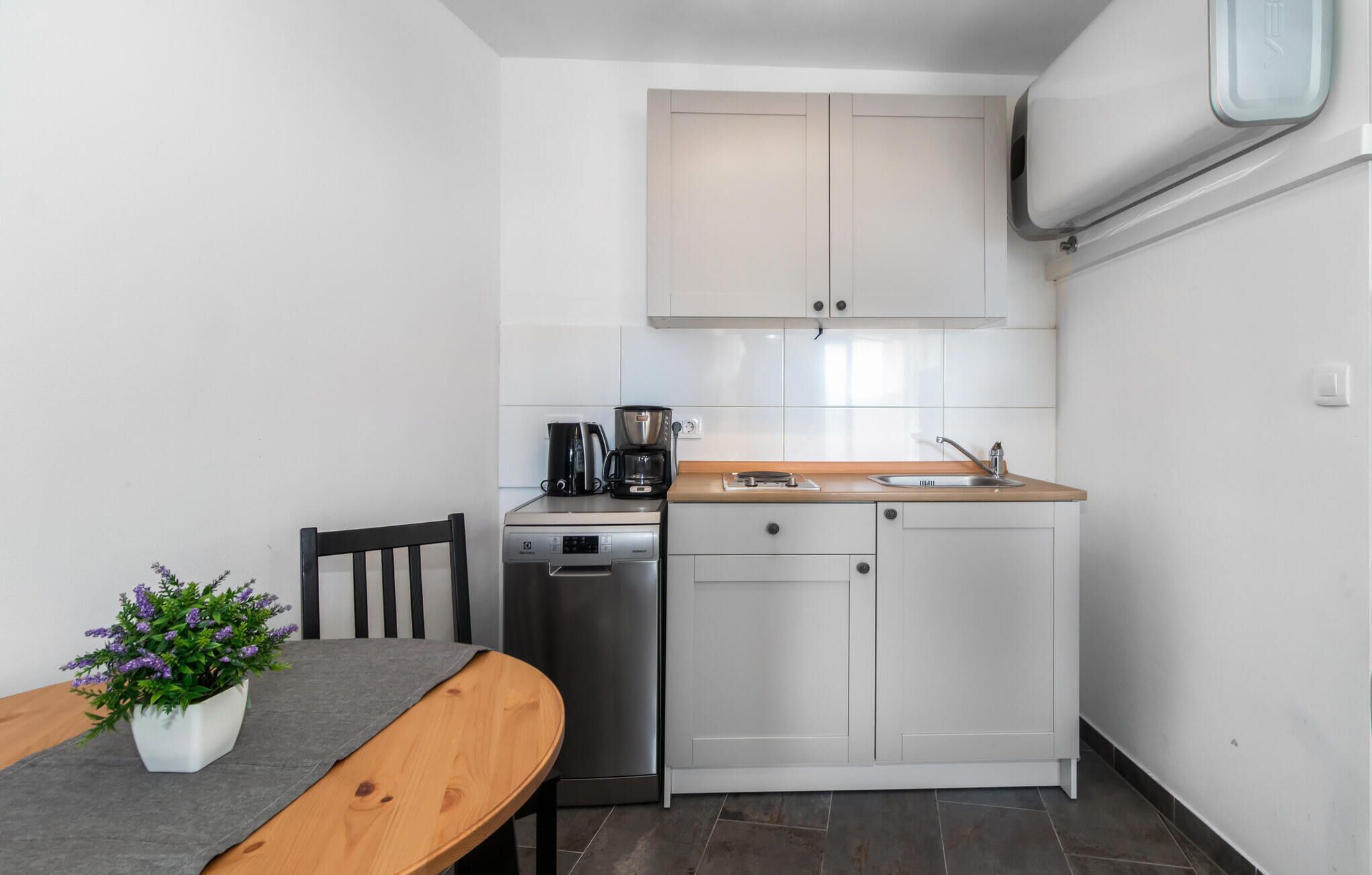 A compact kitchen with white cabinets, a coffee maker, a dishwasher, and a small dining table with a potted plant.