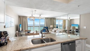 Una cocina moderna con encimera de granito, pileta de acero inoxidable y lavavajillas. Hay una zona de comedor con mesa y sillas, y una sala de estar con sofá y televisor. La habitación tiene ventanas amplias con vista al océano.