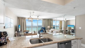 A modern kitchen with a granite countertop, stainless steel sink, and dishwasher. There is a dining area with a table and chairs, and a living room with a sofa and a TV. The room has large windows with a view of the ocean.