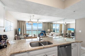 A modern kitchen with a granite countertop, stainless steel sink, and dishwasher. There is a dining area with a table and chairs, and a living room with a sofa and a TV. The room has large windows with a view of the ocean.