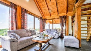 Ein geräumiges Wohnzimmer mit Holzbalkendecke, großen Fenstern und Blick ins Freie.