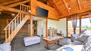 Ein Holzblockhaus-Innenraum mit Treppe, einem Wohnzimmer mit Sofa und Couchtisch sowie einer durchgehenden Tür erkennbaren Küchenzeile.