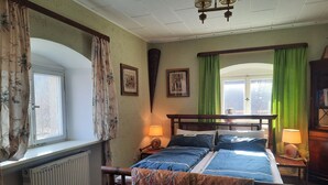 A bedroom with a wooden bed, two bedside tables with lamps, a radiator, a window with curtains, and a green curtain on the window. - Living in a stately home from 1848 in the Fichtelgebirge (Ebnath)