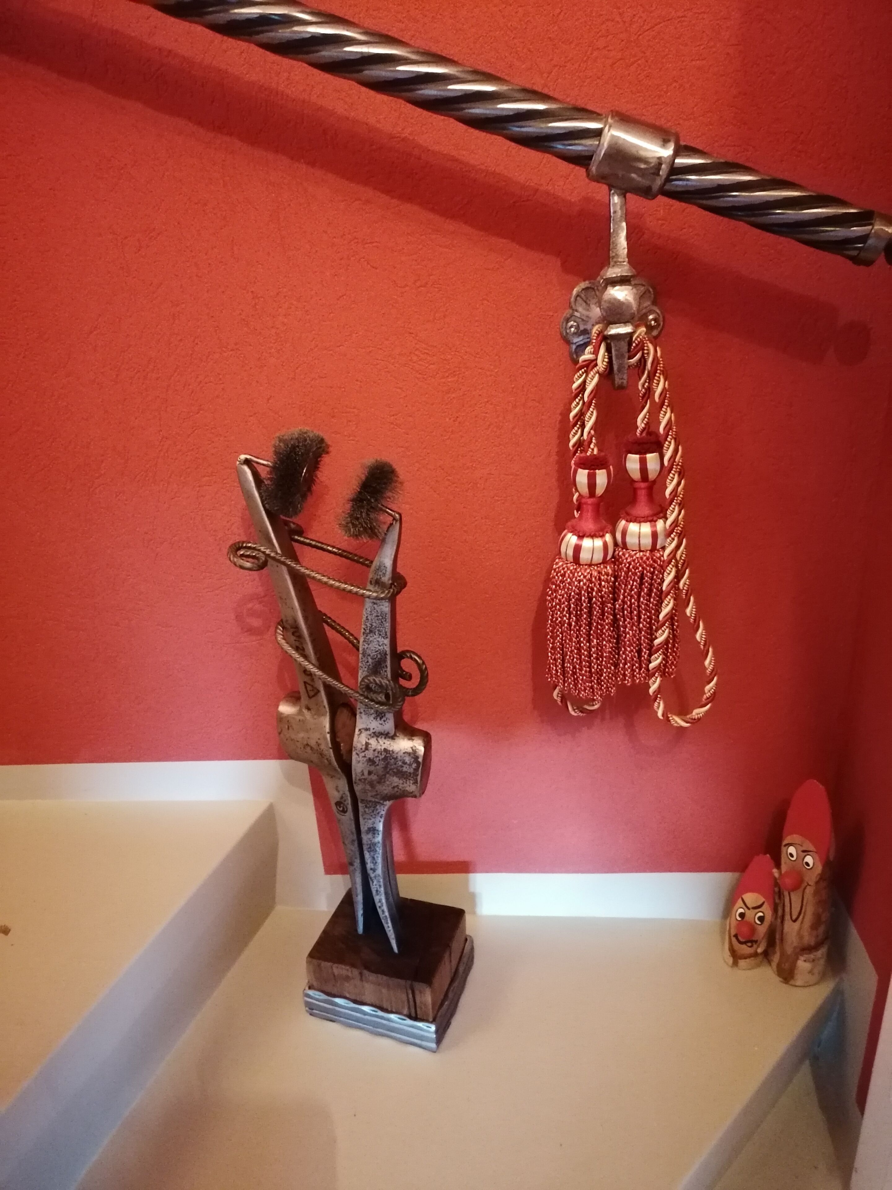 A room with a red wall, a metal sculpture, a rope, and a small shelf with two figurines.
