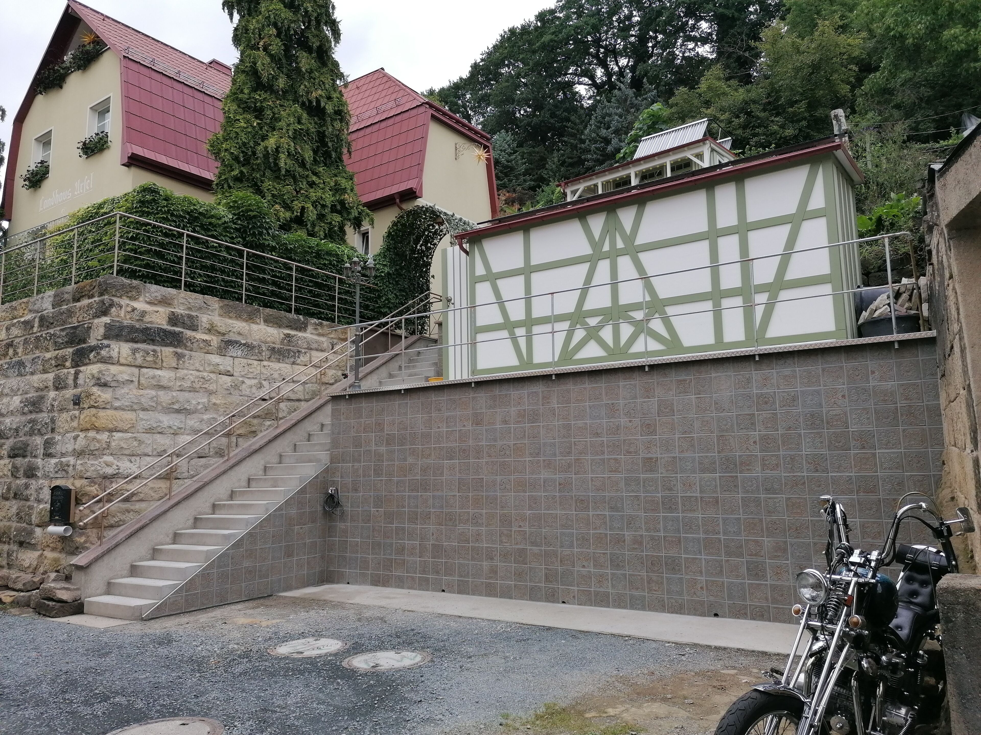 A building with a stone staircase leading to a door, a motorcycle parked nearby, and a building with a green and white facade in the background.