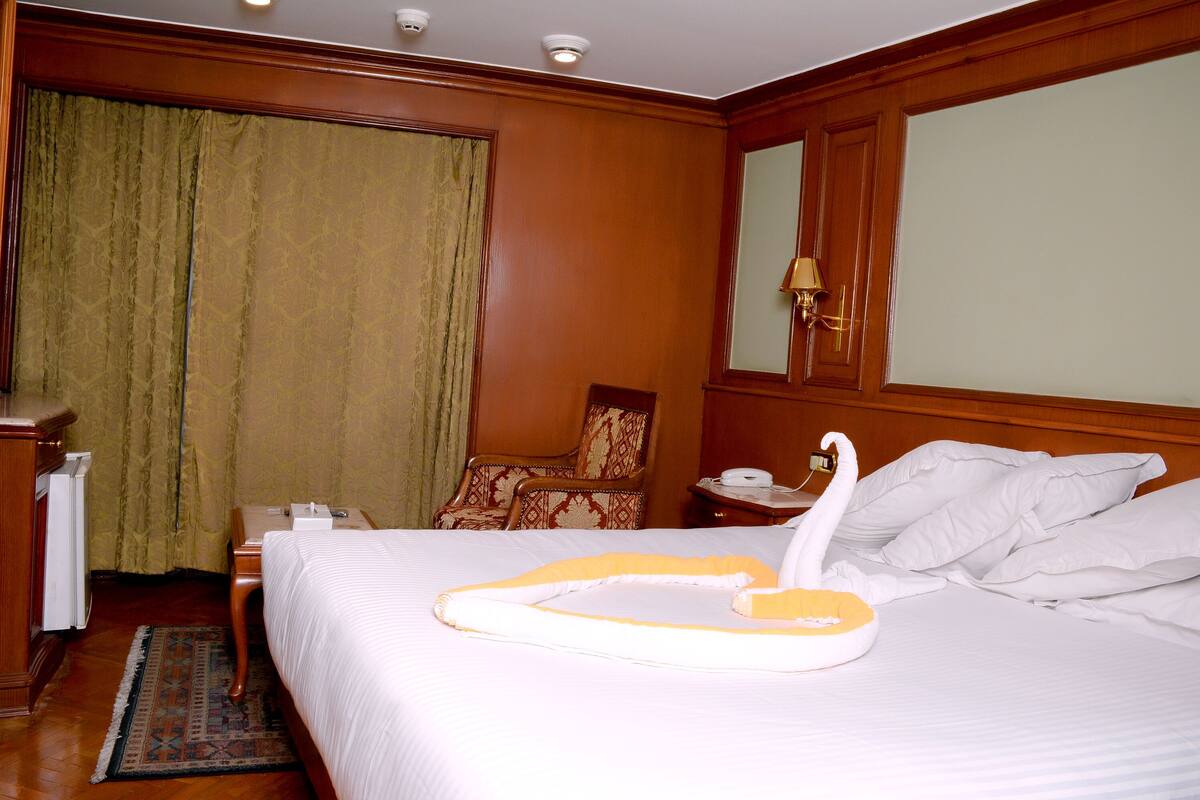 Une chambre d’hôtel avec un grand lit, une tête de lit en bois, une table de chevet avec une lampe, une chaise et un bureau avec un téléphone.