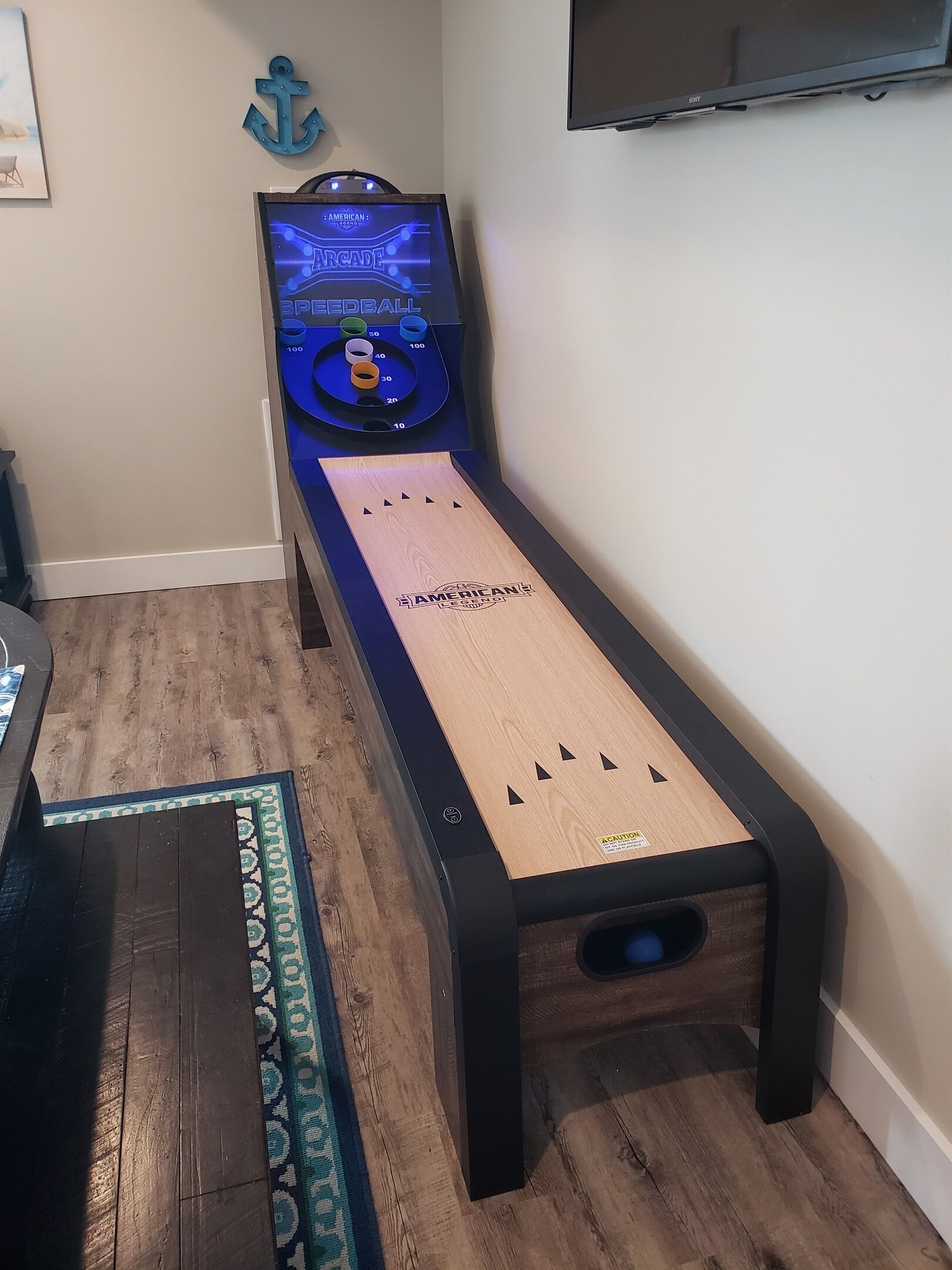 A shuffleboard table with a wooden finish and a blue border, positioned in a room with wooden flooring and a blue rug.