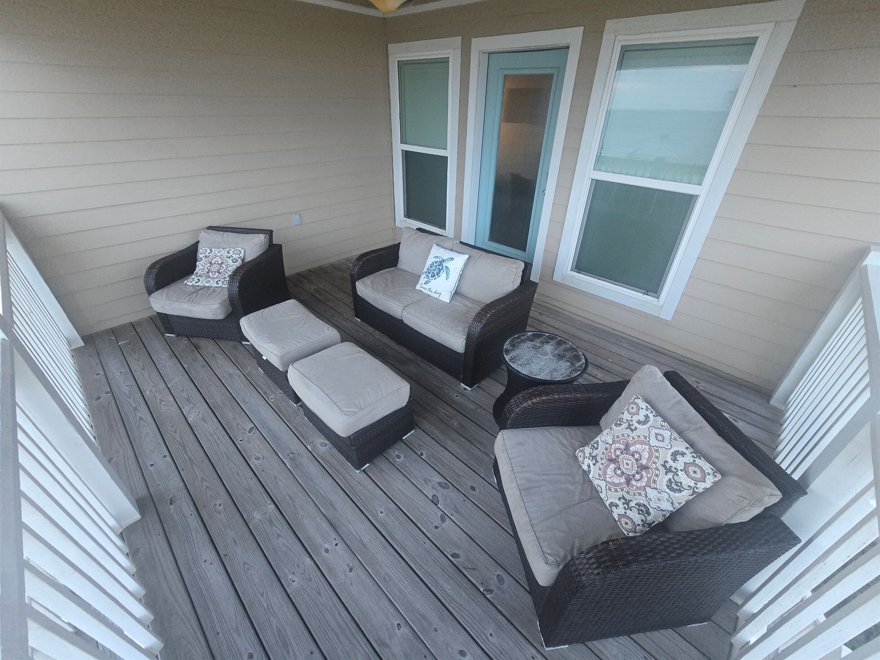 A covered outdoor seating area with wicker furniture and cushions, a small round table, and large windows with blinds.